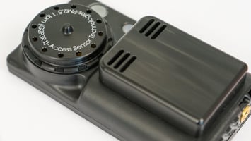 A photograph of an Ultrasonic Personal Air Sampler v2.1 PLUS (a small, black, wearable air sampler) that focuses on the inlet and outlet air flow paths for the time-resolved low-cost particulate matter sensor. 