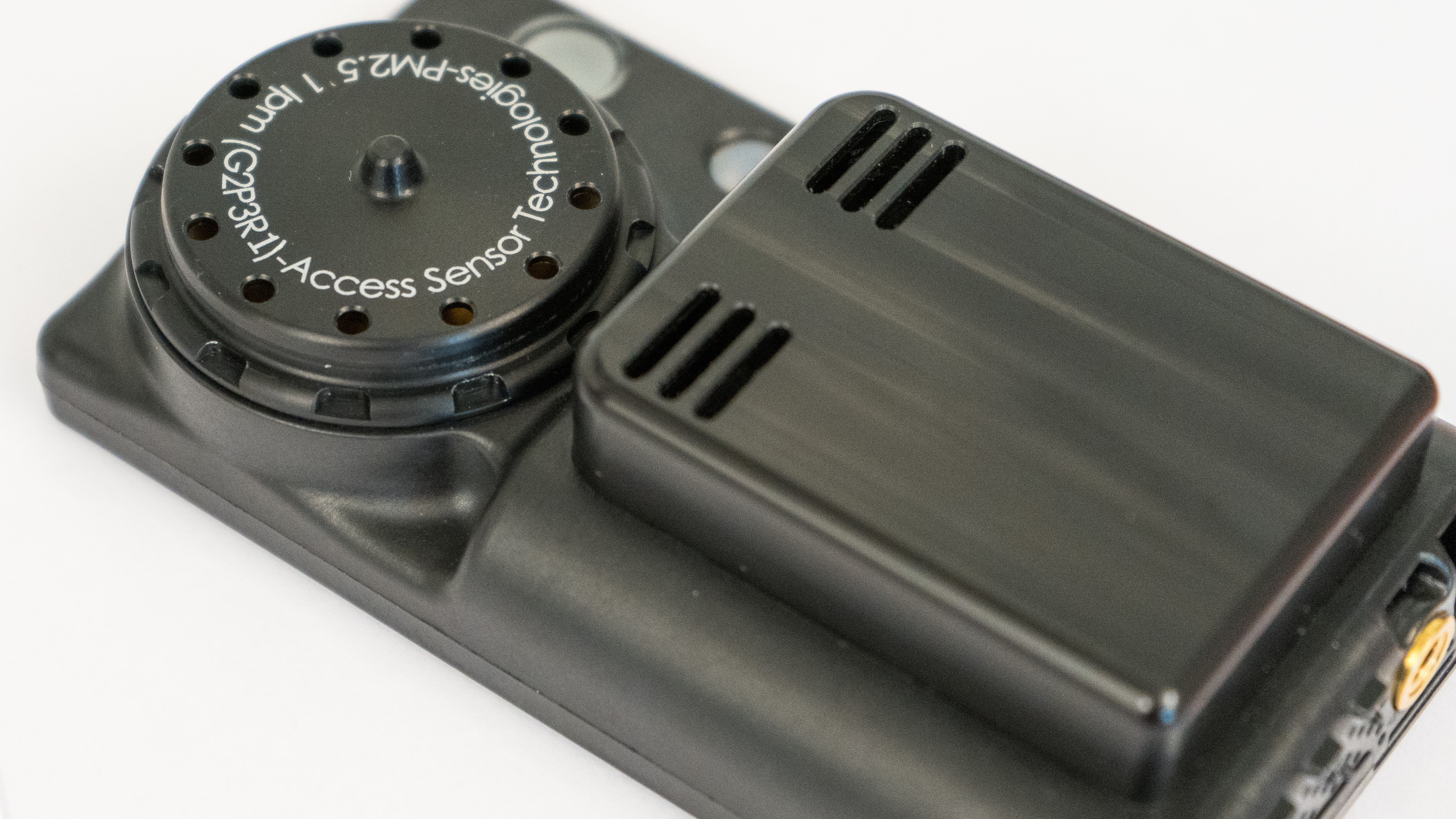 A photograph of an Ultrasonic Personal Air Sampler v2.1 PLUS (a small, black, wearable air sampler) that focuses on the inlet and outlet air flow paths for the time-resolved low-cost particulate matter sensor. 